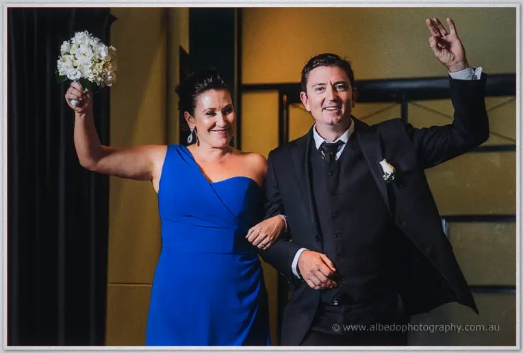 A groomsman waves to the crowd as his partner in blue raises her white posy triumphantly on arrival.