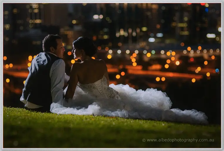 Seated back to back on night grass, Allison's train spilling toward the camera over Perth's orange city bokeh.