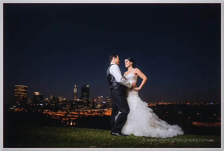 Greg holds Allison around the waist on the hilltop as the full Perth skyline glitters under a deep blue sky.