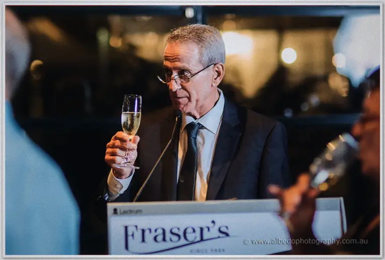 An older gentleman in glasses raises a champagne flute during a toast at the Fraser's podium.
