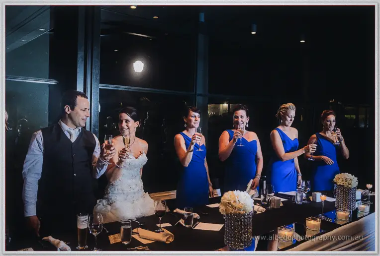 Allison, Greg and the bridesmaids in blue raise champagne flutes together during toasts in the darkened room.
