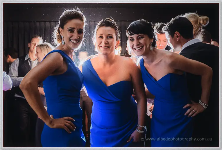 Three bridesmaids in royal blue grin arm in arm on the packed dance floor, flash flare behind them.