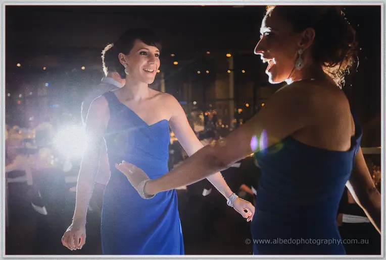 Two bridesmaids in blue dance hand in hand, backlit flare and lens sparkle lighting the busy room.