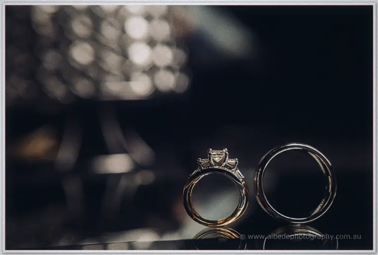 A diamond solitaire ring and Greg's polished band catch the candlelight on a mirror-black table.