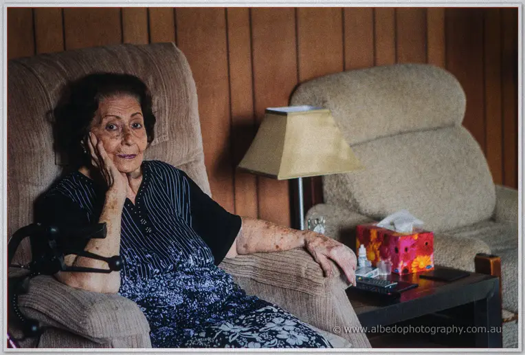 Elderly woman in a dark floral dress rests her cheek on her hand in an armchair by lamplight