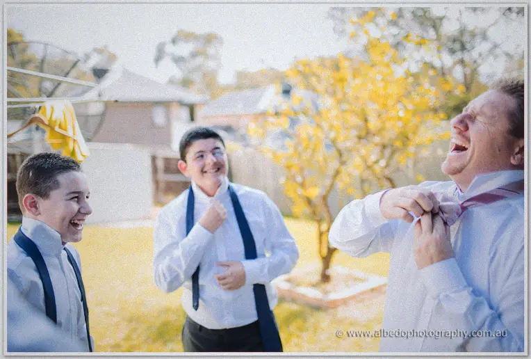 Groom laughs as friends adjust his pink striped tie in a backyard with an autumn-lit tree