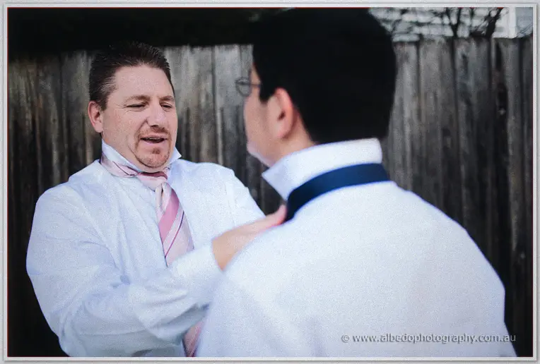 Groomsmen in white shirts help each other with ties and navy braces in a backyard beside a fence