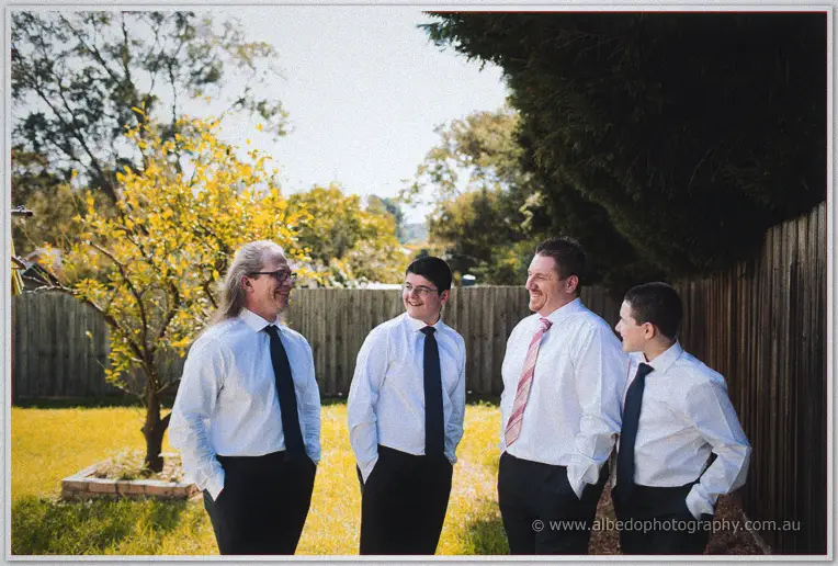 Four men in shirts and navy ties laugh together on a sunlit lawn beside a timber fence