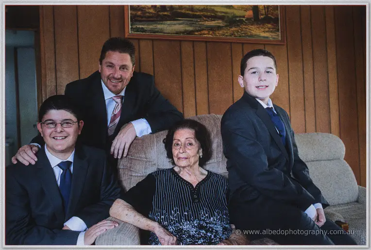 Three generations of Mark's family in dark suits gather around his seated grandmother in a lounge