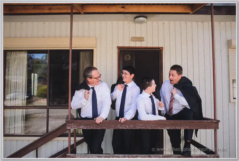 Mark and his groomsmen joke on the veranda of a cream weatherboard house, jackets over shoulders