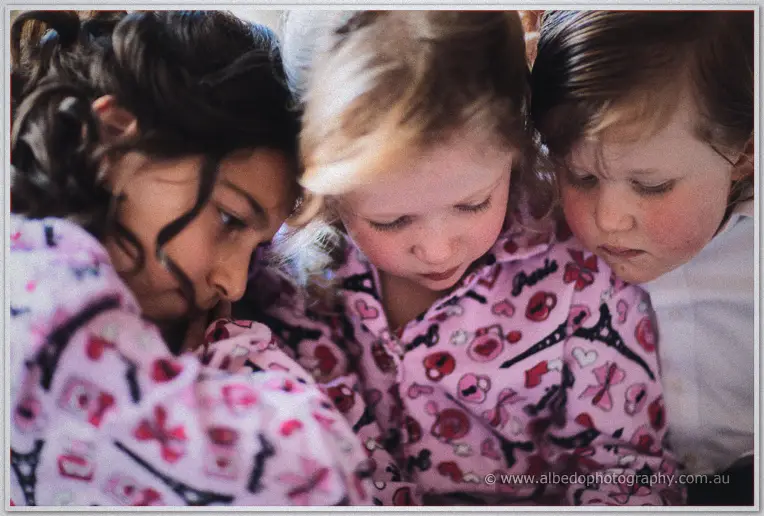 Three children huddle close together, heads bent over something unseen, in pink Paris-print pyjamas