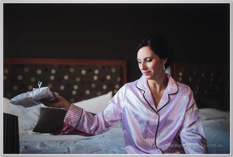 Felicity in striped satin pyjamas sits on a bed holding up a white lace garter in warm room light