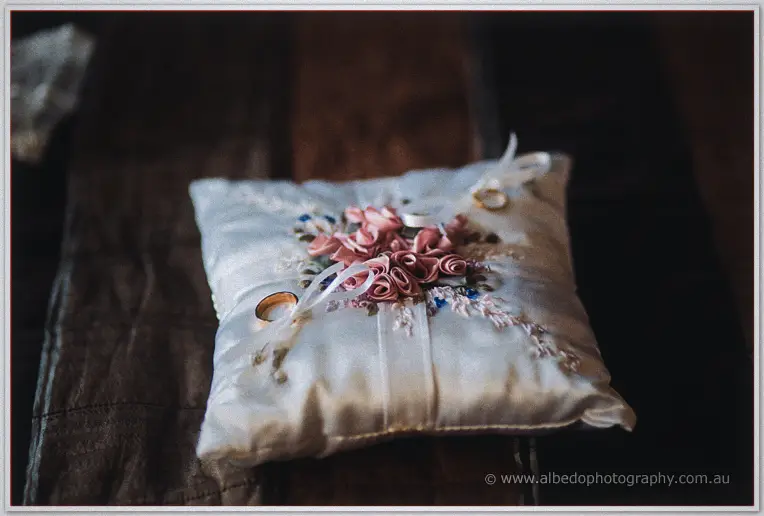 Satin ring pillow with pink ribbon roses holds two gold wedding bands on a dark timber bench