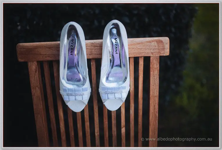 Pair of white peep-toe bridal shoes with jewelled straps hooked over the rail of a wooden garden chair