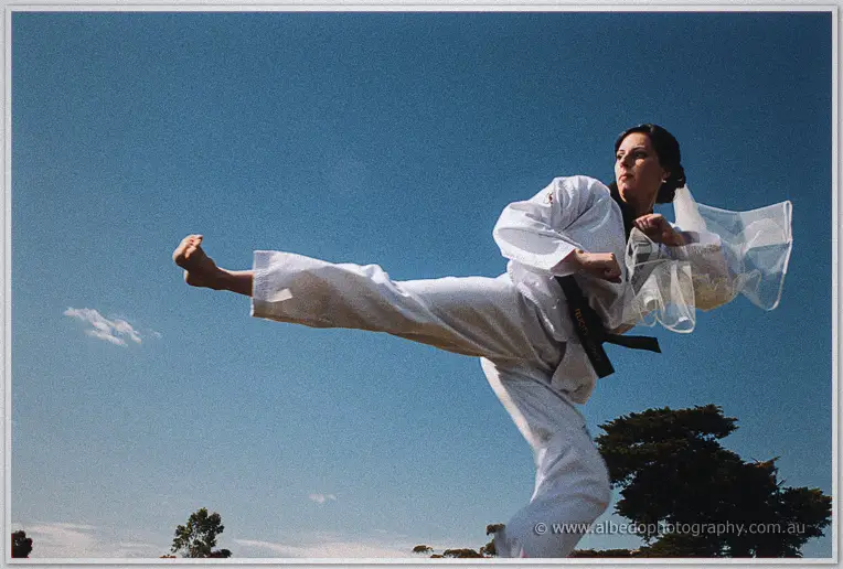 Felicity in a white taekwondo uniform and black belt throws a high kick against a blue sky, veil flying