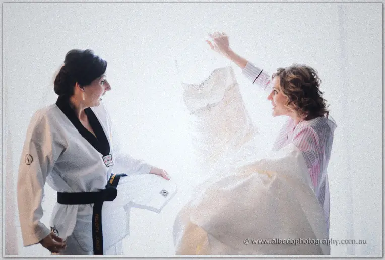 Bridesmaid in a karate uniform and friend in pyjamas laugh while lifting the gown in bright window light