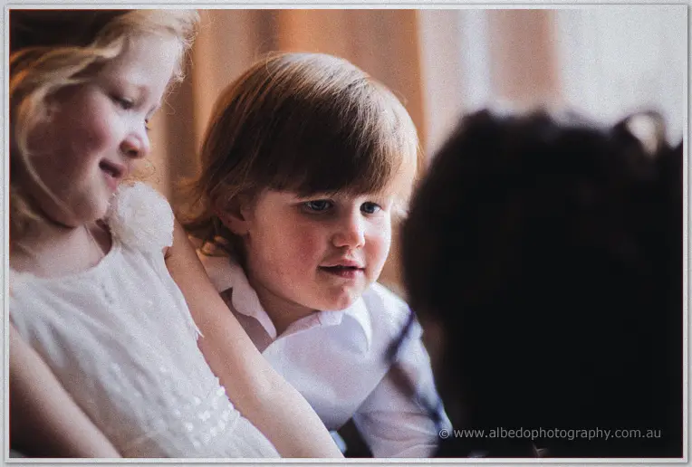 Flower girl in a white dress and young page boy in a white shirt grin together in warm indoor light