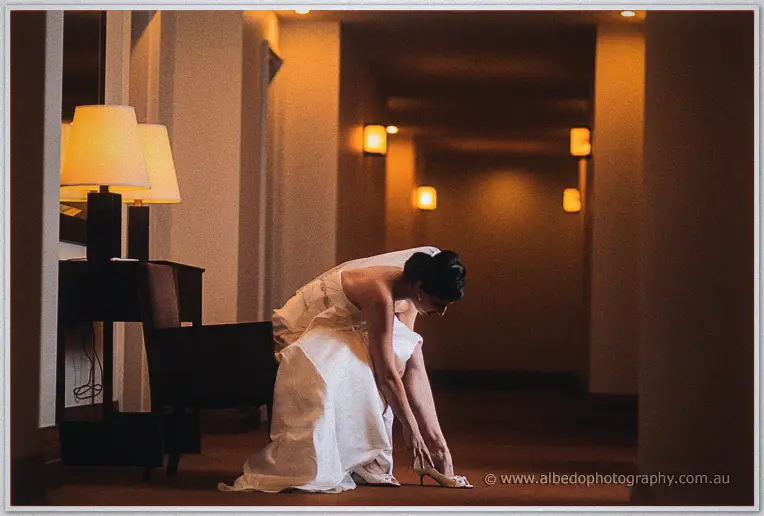 Felicity bends in her gown to fasten a shoe in a warmly lit hotel hallway lined with glowing lamps