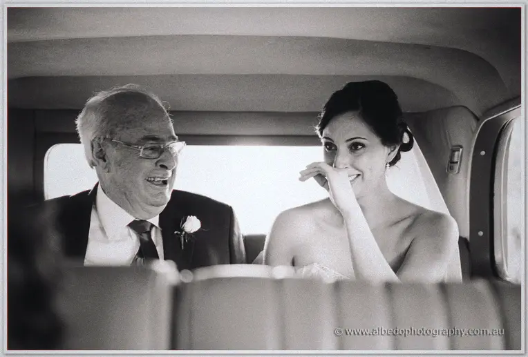 Felicity laughs beside her suited father in the back seat of the bridal car in soft black and white