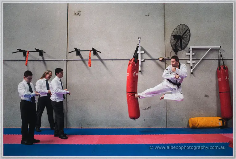 Mark in his suit and taekwondo trousers leaps mid-air with a flying kick at a red punching bag
