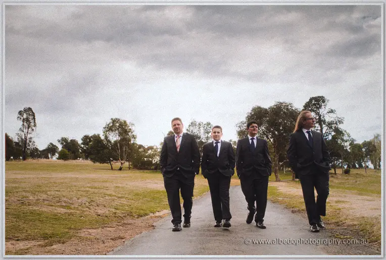 Mark and his groomsmen stroll down a country road in dark suits beneath a heavy grey sky