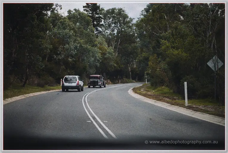 A vintage bridal car follows a white car down a tree-lined country road under an overcast sky