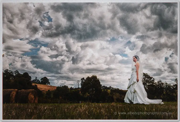 Veiled bride in her flowing gown pauses on a wide paddock beneath a dramatic stormy sky