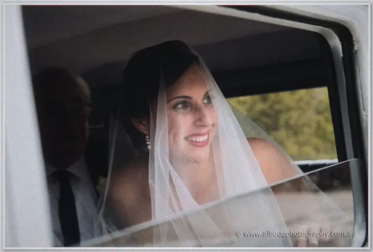 Veiled Felicity smiles brightly from the bridal car window with her father seated behind her