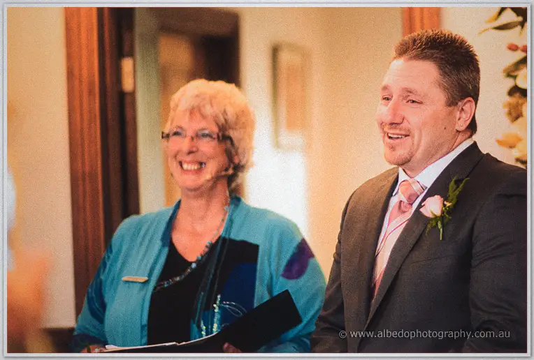 Beaming groom in a grey suit and pink rose boutonniere stands with the smiling celebrant indoors
