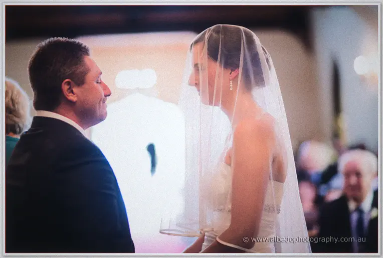 Veiled Felicity and Mark face each other at the chapel altar in soft, hazy window light
