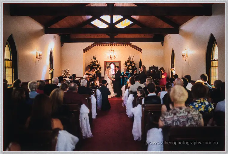 Wide view of the couple exchanging vows in the small timber-beamed chapel before seated guests