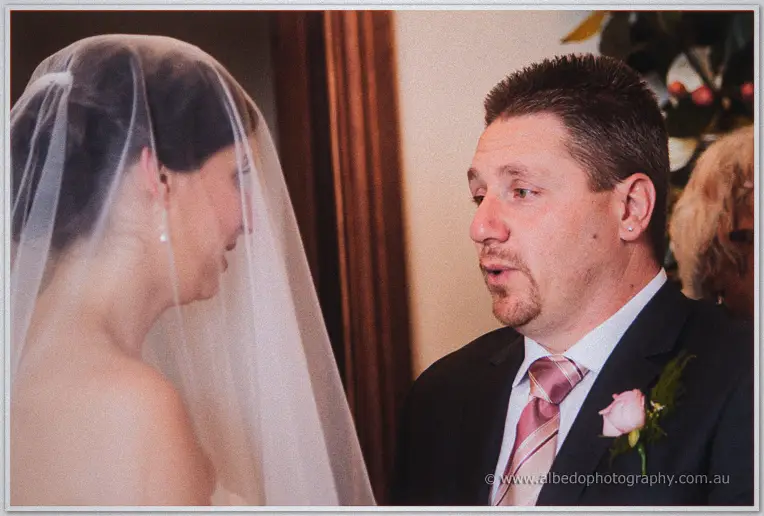 Mark speaks earnestly to veiled Felicity before him, his pink rose boutonniere catching the light