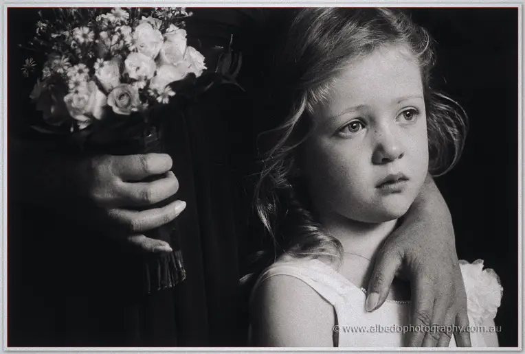 Black-and-white close-up of a young flower girl gazing away, a hand resting on her shoulder