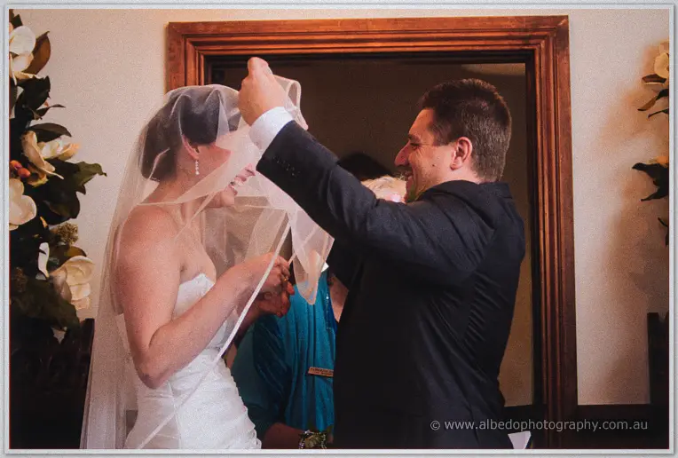 Mark grins as he lifts Felicity's veil back in a warmly lit room with white floral arrangements