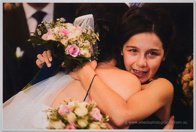 Young girl beams over Felicity's shoulder as they embrace, pastel rose bouquet in hand