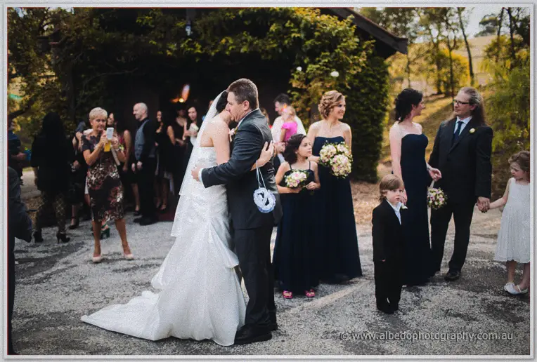 Felicity and Mark embrace outside as guests look on in the tree-lined grounds of Inglewood Estate.