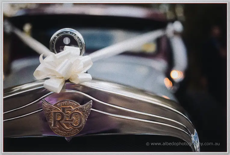 Cream ribbon bow and winged REO radiator badge gleam on the bonnet of the maroon vintage bridal car