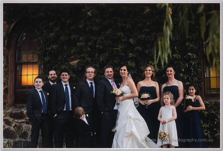 The bridal party poses before the ivy-draped stone chapel wall, warm light glowing in its window.