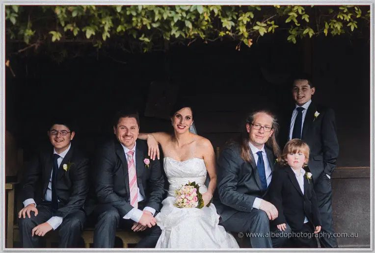 Felicity sits with family on a shaded bench, arm around Mark, all smiling for the camera.