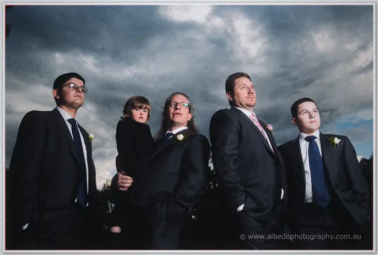 Mark and his groomsmen stand moody under a stormy sky, one holding the young pageboy.