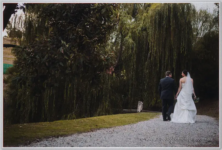 Seen from behind, the couple wanders a gravel path beneath weeping willows.