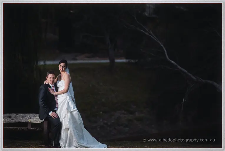 Mark sits on a rustic bench with Felicity standing close in deep twilight shade.