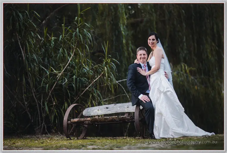 Felicity leans on Mark as he sits on an old wheelbarrow seat under willows.