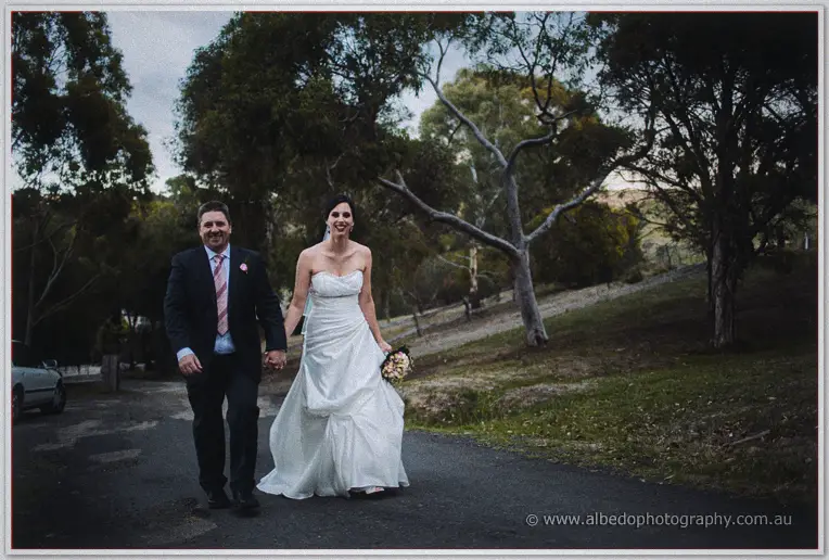 The newlyweds stroll hand in hand down a tree-lined gravel road at dusk.