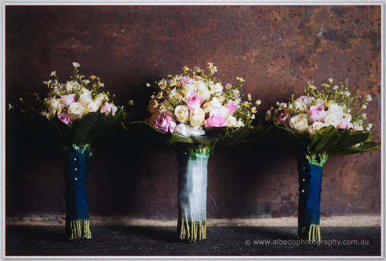 Three pastel rose and daisy posies with navy ribboned stems lined up against a rustic rust-coloured wall