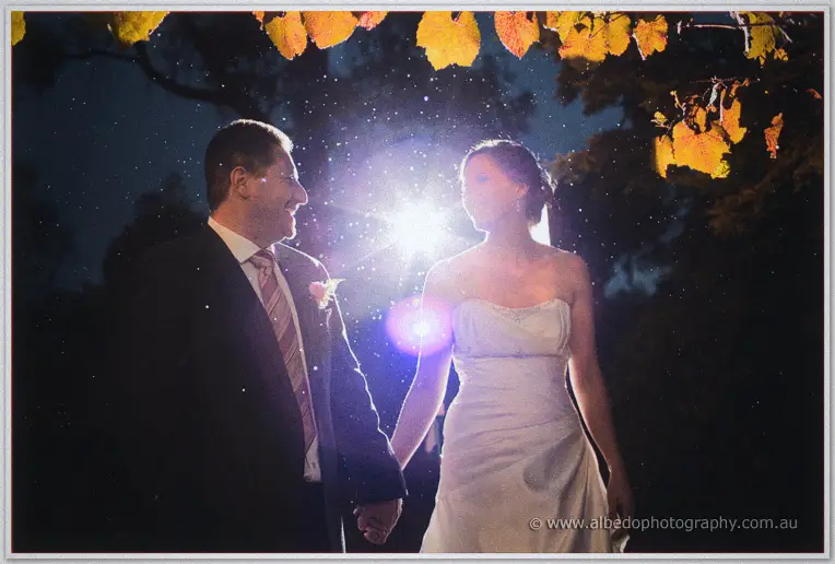 Camera flash flares between the laughing newlyweds beneath golden autumn leaves.