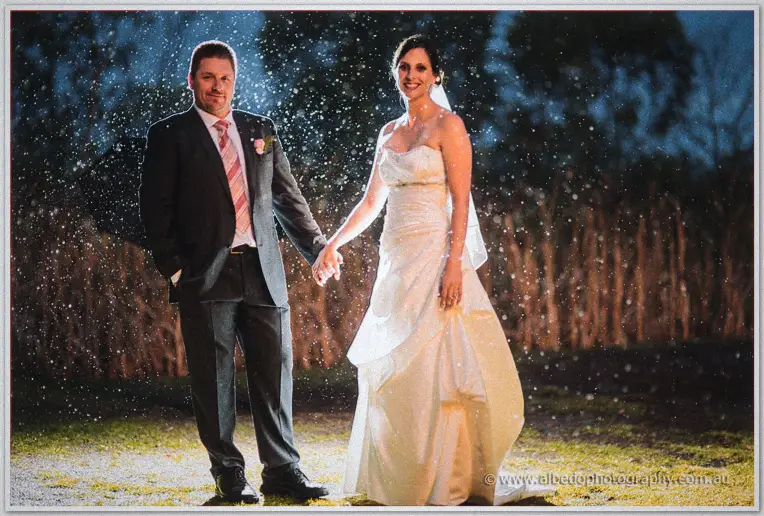 The couple holds hands through drifting rain, lit against the dark bushland.