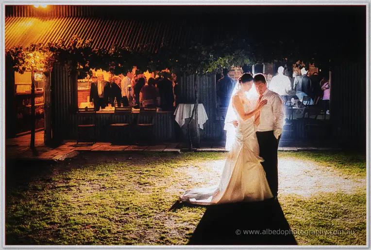 The couple stands close on the lawn as reception guests glow behind glass doors.