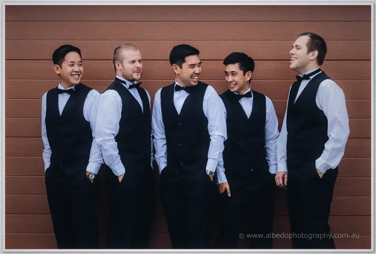 Chris and his groomsmen laugh together in black vests and bow ties against a terracotta wall.