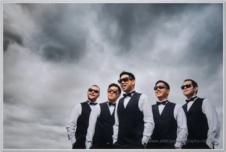 Groomsmen in sunglasses smile beneath a dramatic clouded sky, Chris grinning at the centre.